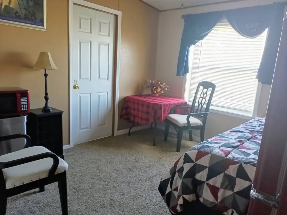 The image showcases a warmly lit bedroom with a patterned bed, a small dining table covered in a red checkered tablecloth, a cozy lamp, and a window framed by curtains that lets in soft natural light. The scene feels inviting and homey, perfect for relaxing or enjoying a quiet meal while gazing outside.