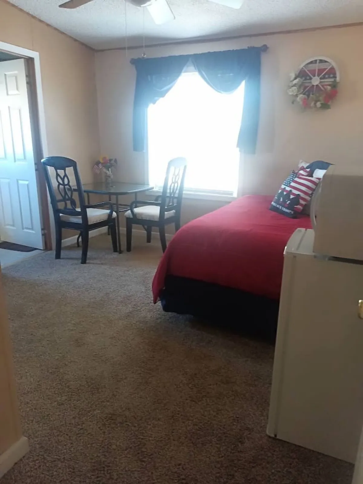 An inviting living space with a bed adorned in red bedding, a dining table with chairs, and a window streaming natural light through airy curtains. The refrigerator is visible, suggesting the space is both functional and warm, ideal for relaxing or enjoying a meal.
