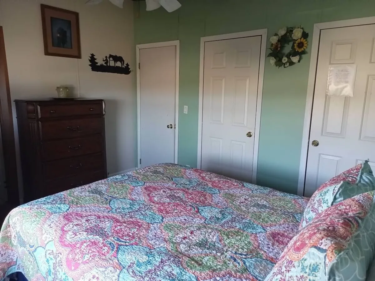 This interior photo features a cozy bedroom with a colorful quilt, a wooden dresser, decorative wall art, and doorways that suggest privacy and comfort. Sunlight filters gently into the room, emphasizing a restful, welcoming atmosphere.