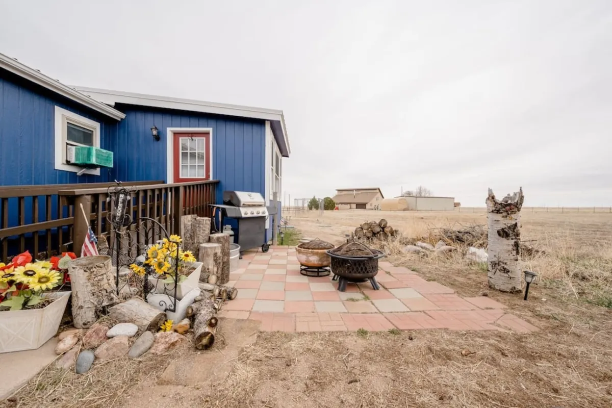 A welcoming outdoor patio area showcasing blue house siding, a grill, a fire pit, flower pots, and a stone path. The setting invites outdoor cooking, gathering around the fire, and relaxed evenings under the open sky.