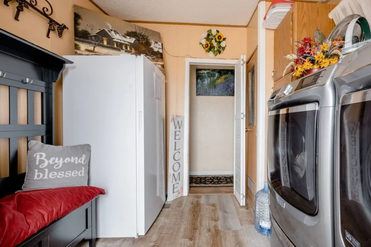 A cozy entry area with a visible fridge, washer and dryer, a welcoming sign, and a comfortable bench with a cushion. The space is decorated with wall art and has a homey, inviting atmosphere, immediately setting a welcoming tone.