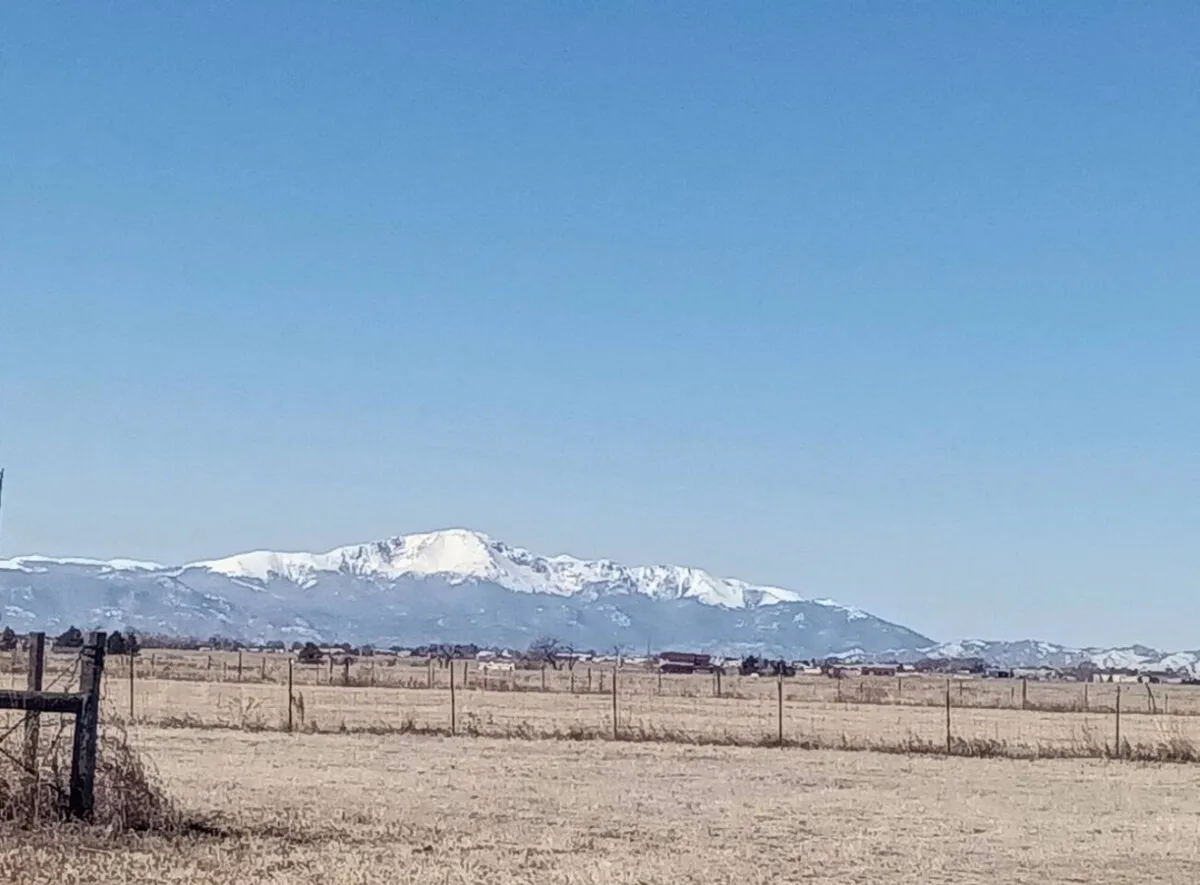 A panoramic view of open fields stretching toward majestic, snow-capped mountains in the distance. A rustic fence runs through the landscape, emphasizing the vastness and rural charm of the property. The lighting is soft, conveying a tranquil, expansive atmosphere.