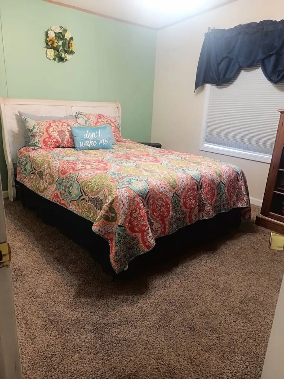 A bright and inviting bedroom with a colorful bedspread, a decorative pillow with text, and floral wall art. The natural light streaming through the window highlights the room’s cheerful, restful ambiance.