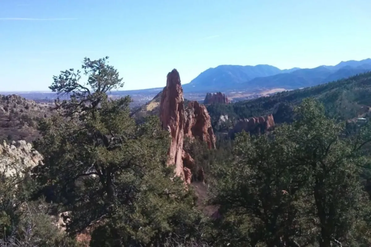 A rugged, natural setting with tall rock formations standing against a backdrop of mountains and dense greenery. The area is bathed in natural light and suggests an environment ideal for hiking and exploring unique geological features, reminiscent of the nearby Paint Mines.