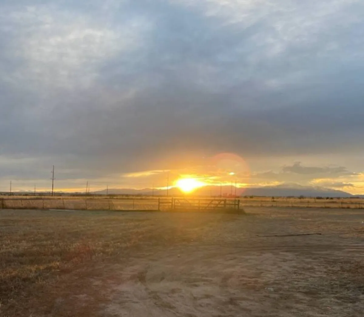 A tranquil exterior shot featuring a vivid sunset over the mountains, a fence line, and open land. The expansive sky and fading light evoke the peaceful transition from day to evening, creating the perfect setting for stargazing or storytelling outdoors.