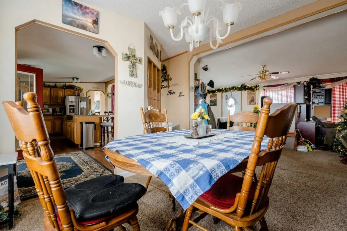 A cozy dining area with a large wooden dining table, a chandelier overhead, and the entrance to the kitchen visible in the background. Decorative wall items add a touch of homeliness. The setting is perfect for group meals, celebrations, and lively conversations.