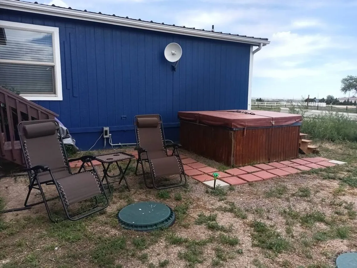An inviting patio scene featuring a hot tub, lounge chairs, and the blue exterior wall of the house. The setup encourages outdoor leisure and socializing, with the hot tub as the centerpiece for both relaxation and gatherings. The space is bathed in soft evening light, suggesting comfort and year-round usability.