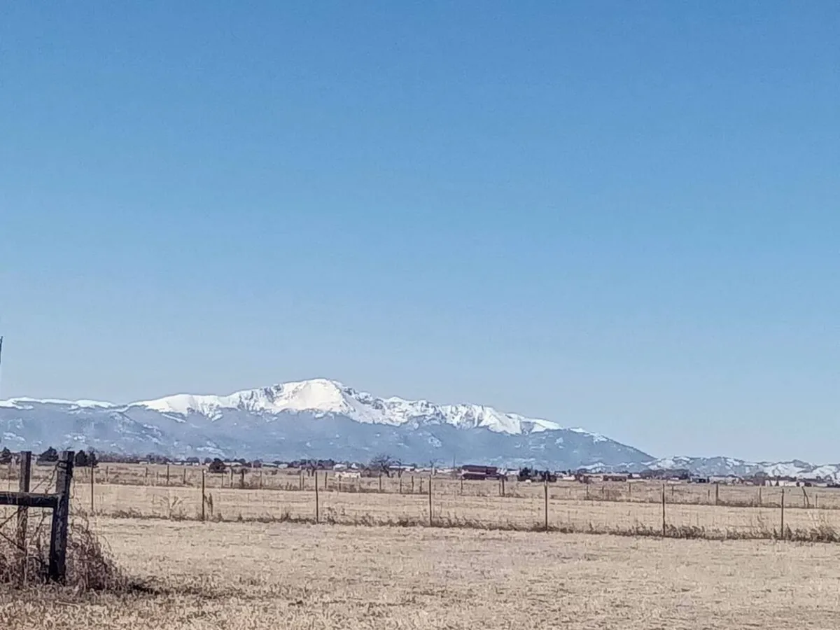 A dramatic landscape featuring snow-capped mountains rising behind a vast open field, with a rustic fence line stretching into the distance. The gentle light hints at sunset, casting warm tones across the land. The scene suggests ample space for hiking, wildlife observation, and taking in the grandeur of the Colorado outdoors.