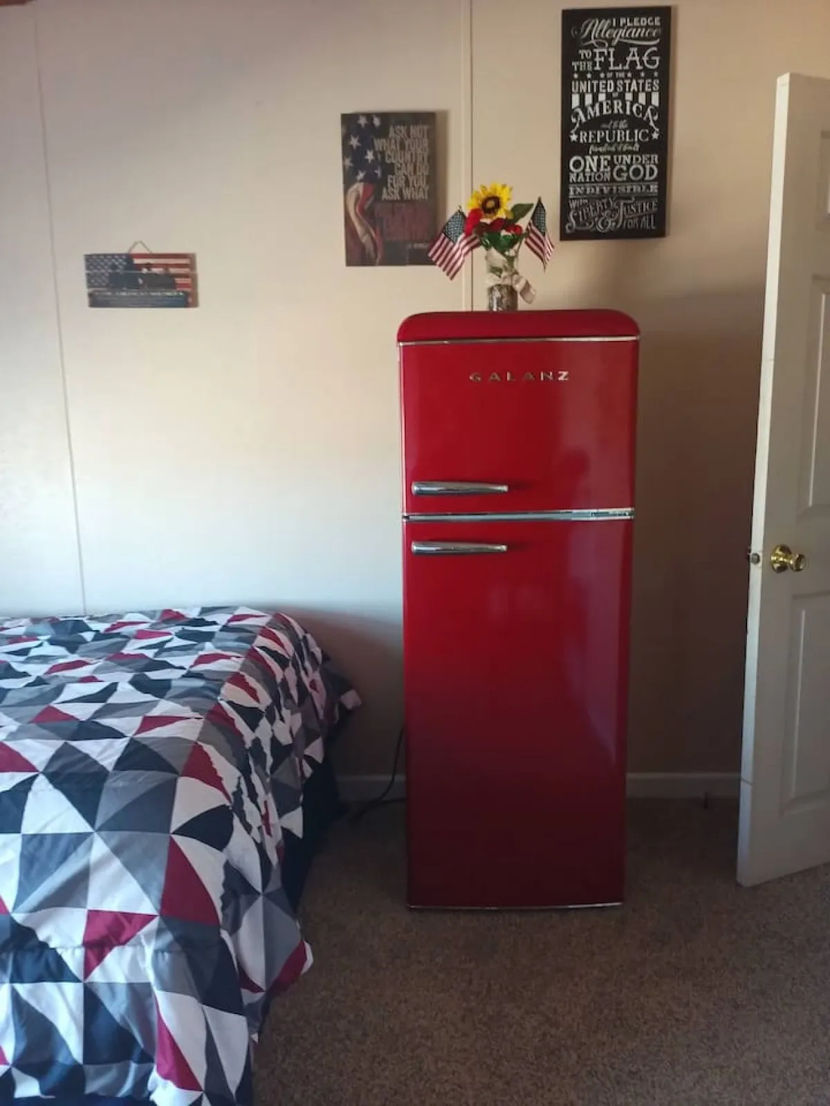 A homey interior with a red refrigerator, quilted bedspread, decorative signs, and a flower arrangement. The layout suggests a multifunctional space where cooking, relaxing, and socializing naturally blend together.