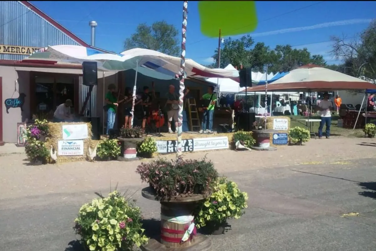 A lively outdoor scene with a stage featuring musicians, colorful flower pots lining the area, and bustling market stalls in the background. People are gathered, enjoying live music and socializing in a community-oriented setting.
