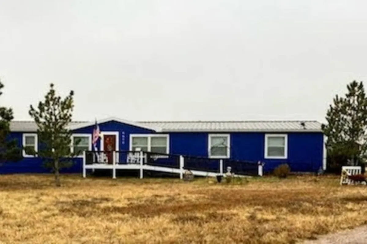 A calm, inviting shot of the property’s blue exterior with white porch railings surrounded by mature trees. The scene suggests a peaceful environment for enjoying the outdoors, morning coffee, or quiet conversation as the sun sets.